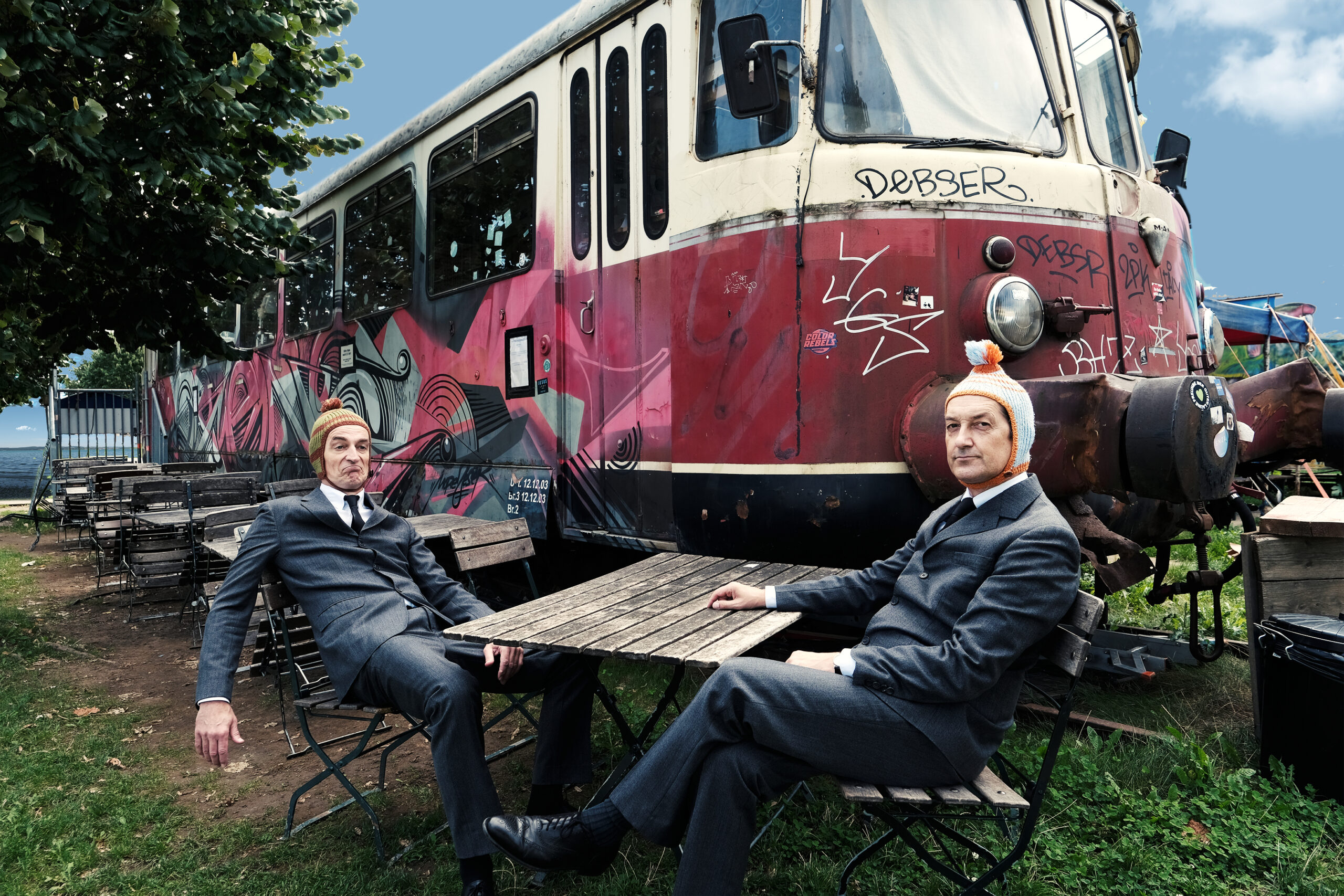 Komikerduo Ulan & Bator sitzen an einem Gartentisch vor einem ausrangierten Straßenbahnwagen.